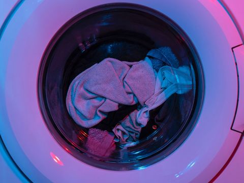 Un primo piano del cestello di una lavatrice con vari indumenti, tra cui un asciugamano grigio chiaro e indumenti scuri. La scena è illuminata con luci rosa e blu, creando un effetto colorato.