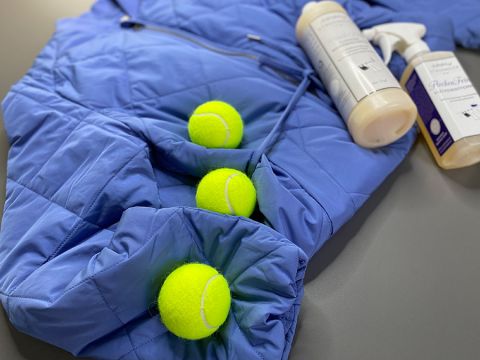 Distesa c'è una giacca a vento blu trapuntata, nelle cui pieghe sono parzialmente infilate tre palline da tennis gialle. Accanto, su una superficie grigia, ci sono due flaconi di detergenti.