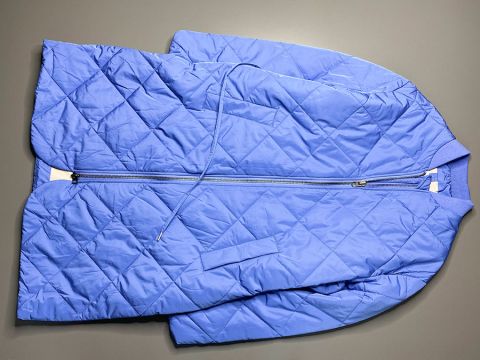 Una giacca trapuntata blu con zip frontale è distesa su una superficie grigia. La giacca ha un motivo a rombi e un cordoncino in vita.