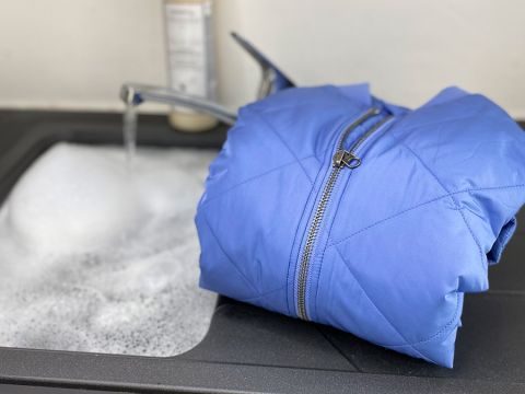 Una giacca blu è appoggiata sul bordo di un lavandino pieno di acqua saponata, sullo sfondo un rubinetto è aperto.