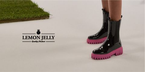 Persona con stivali neri lucidi con suole rosa su sfondo bianco, accanto a una macchia di erba verde. A sinistra c'è il testo "Lemon Jelly Quality Product".
