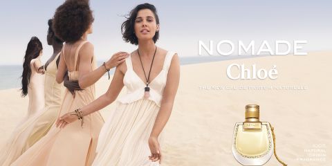 Quattro donne camminano attraverso un paesaggio desertico in abiti ampi. A destra si vedono il testo "Nomade Chloé" e una bottiglia di profumo che pubblicizza la fragranza. La scena trasmette una sensazione di libertà ed eleganza naturale.