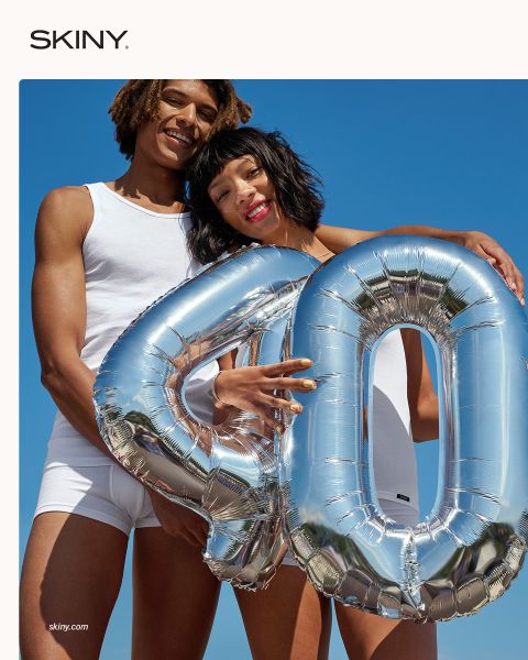 Due persone in biancheria intima bianca sorridono tenendo un palloncino argentato con il numero "40" davanti a un cielo azzurro brillante.