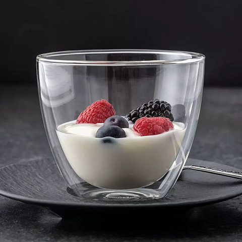 Un bicchiere a doppia parete è riempito con yogurt cremoso e guarnito con lamponi, more e mirtilli freschi. Il bicchiere è posizionato su un piattino nero con un cucchiaio accanto, su uno sfondo scuro.