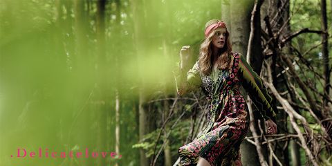Una donna con un vestito a motivi colorati e una fascia rossa per capelli si trova in mezzo a una lussureggiante foresta verde. Nell'angolo in basso a sinistra, le parole "Delicatelove" sono scritte in rosa. Lo sfondo è sfocato con foglie verdi.