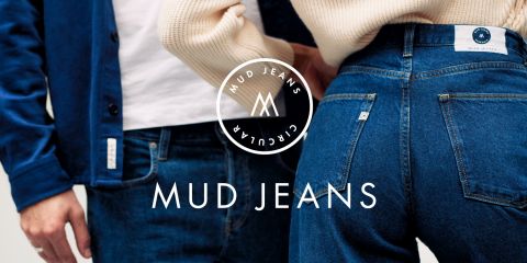 Due persone sono in piedi vicine, indossando jeans blu Mud. La persona a sinistra indossa una giacca e la persona a destra mostra la parte posteriore dei jeans. Il logo e il testo di Mud Jeans sono chiaramente visibili.