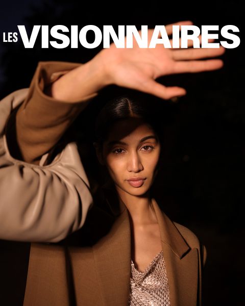 Una persona con un cappotto marrone alza una mano per coprire parzialmente il viso. Si trova di fronte a uno sfondo scuro. Nella parte superiore dell'immagine, le parole "Les Visionnaires" sono scritte in grassetto bianco.
