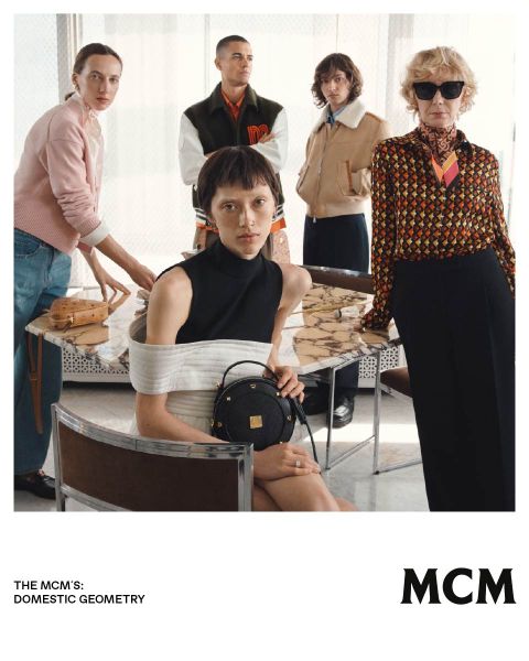 Cinque persone eleganti posano in un interno attorno a un tavolo di marmo, presentando borse e moda MCM in un ambiente moderno ed elegante.