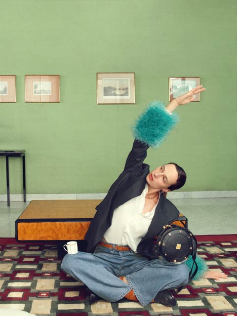 Una persona in jeans e un blazer con polsini blu e pelosi posa con il braccio alzato in una stanza elegante con pareti verdi, opere d'arte incorniciate e un tappeto a motivi.