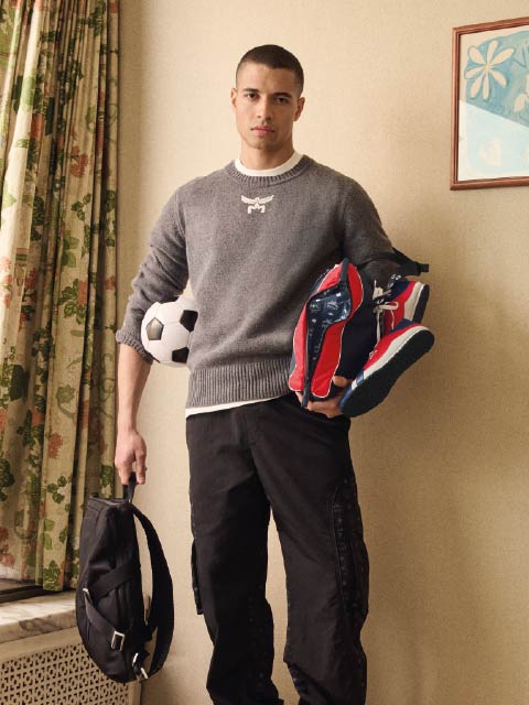Un giovane uomo con un maglione grigio tiene in mano uno zaino nero e delle scarpe da ginnastica, un pallone da calcio sotto il braccio, e si trova all'interno vicino a tende floreali.