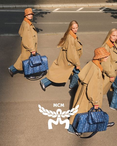 Tre donne con cappotti marrone chiaro e cappelli arancioni camminano per una strada, portando borse MCM blu con un logo MCM bianco in primo piano. L'immagine ha un effetto specchio che fa apparire la scena duplicata.
