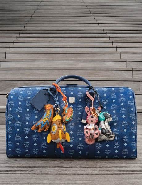 Una borsa firmata MCM blu con motivo logo bianco è appoggiata su gradini di legno decorati con colorati portachiavi a forma di animali e oggetti, tra cui un razzo, un coniglio e un orso.