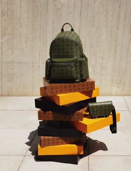 Uno zaino di design verde e una borsa a tracolla abbinata sono appoggiati su una pila di scatole arancioni e marroni davanti a una parete strutturata beige e un pavimento piastrellato.