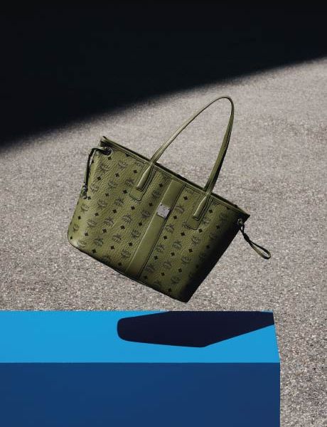 Una borsa verde di design con un discreto motivo monogrammato è appoggiata su una superficie grigia, con un oggetto geometrico blu in primo piano e un'ombra proiettata sullo sfondo.