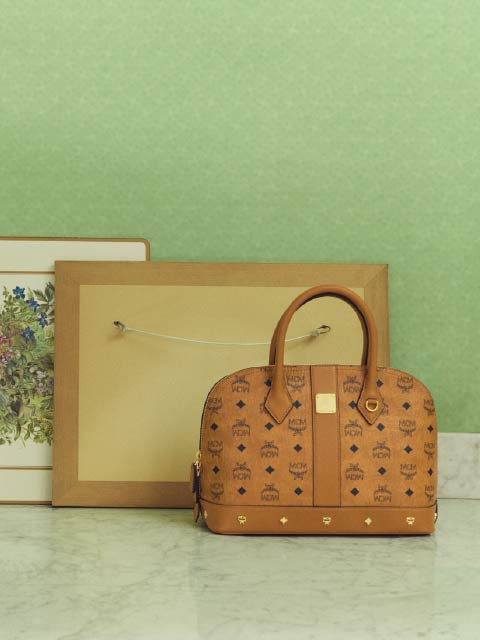 Una borsa firmata marrone giace su una superficie di marmo davanti a due quadri incorniciati, di fronte a una parete verde chiaro.