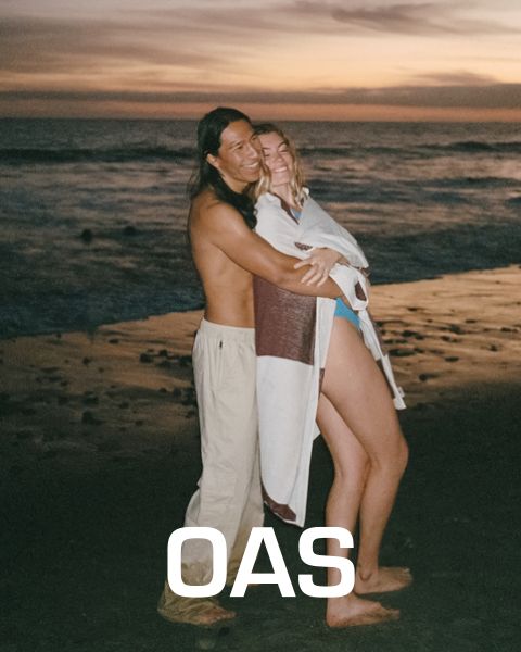 Una coppia si abbraccia al tramonto su una spiaggia. L'uomo è a torso nudo e indossa pantaloni leggeri, mentre la donna è avvolta in una sciarpa bianco-marrone. Sullo sfondo si vedono le onde del mare, sotto c'è il testo "OAS".
