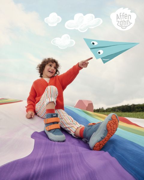 Un bambino con un vivace maglione arancione e pantaloni a righe è seduto su una superficie colorata e indica un aeroplanino di carta in stile fumetto nel cielo. Sorride ed è circondato da nuvole soffici. Nell'angolo c'è scritto "Affenzahn".