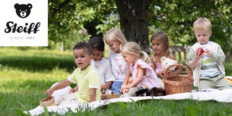Un gruppo di bambini è seduto su una coperta all'aperto, circondato da alberi. Stanno giocando con cesti e peluche. Il logo Steiff si trova nell'angolo in alto a sinistra.
