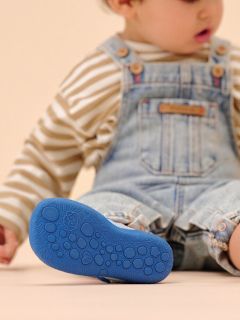 Un bambino piccolo con una camicia a righe e una salopette di jeans è seduto con una gamba all’aperto e mostra la suola blu strutturata della sua scarpa.