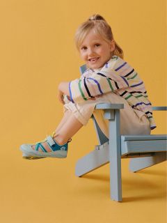 Bambino sorridente con camicia a righe e pantaloni chiari seduto di lato su una sedia di legno blu davanti a uno sfondo giallo.