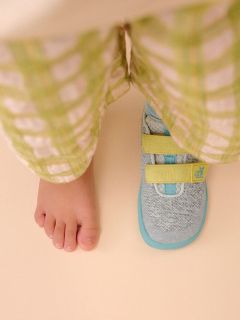 Un piede nudo di un bambino accanto a una scarpa da ginnastica con cinghie gialle, entrambi visibili sotto un paio di pantaloni a quadri verde chiaro su un pavimento beige.