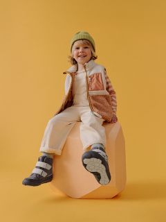 Un bambino sorridente con un cappello verde, una camicia a righe e un gilet è seduto su un blocco color pesca davanti a uno sfondo giallo, indossando stivali e pantaloni chiari.