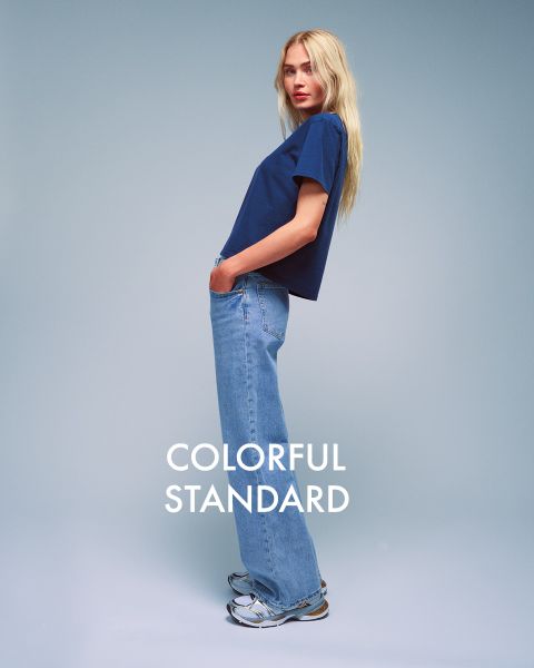 Una persona con lunghi capelli biondi è di profilo su uno sfondo blu. Indossa una maglietta blu scuro, jeans azzurri e scarpe da ginnastica. Il testo "COLORFUL STANDARD" è visualizzato in bianco.