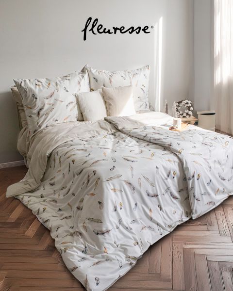 Una camera da letto accogliente con un letto ben fatto con lenzuola bianche con motivo di uccelli. Due cuscini decorativi sono appoggiati alla testiera. La luce del sole filtra attraverso tende trasparenti, proiettando una luce soffusa sul pavimento in legno chiaro.