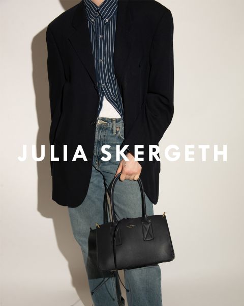 Persona in un blazer nero sopra una camicia a righe e jeans blu, che tiene una borsa nera. Il testo "JULIA SKERGETH" è chiaramente visibile sovrapposto all'immagine.