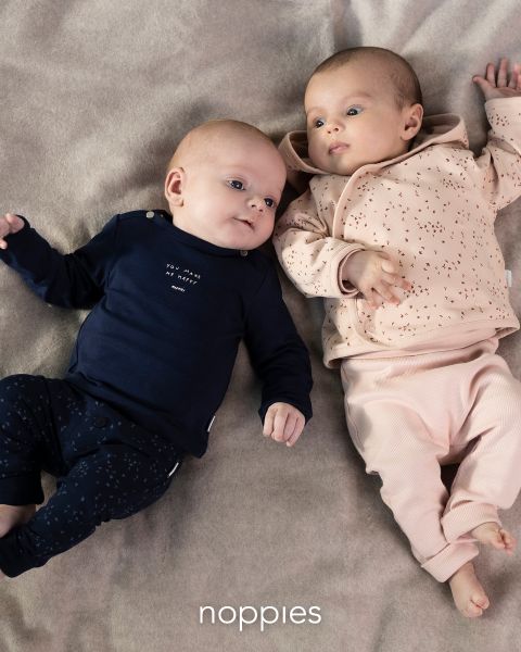 Due neonati sono sdraiati su una coperta morbida. Il neonato a sinistra indossa un completo blu navy con un motivo bianco, mentre il neonato a destra indossa un completo color pesca con macchioline più scure. Nella parte inferiore dell'immagine appare la parola "Noppies".