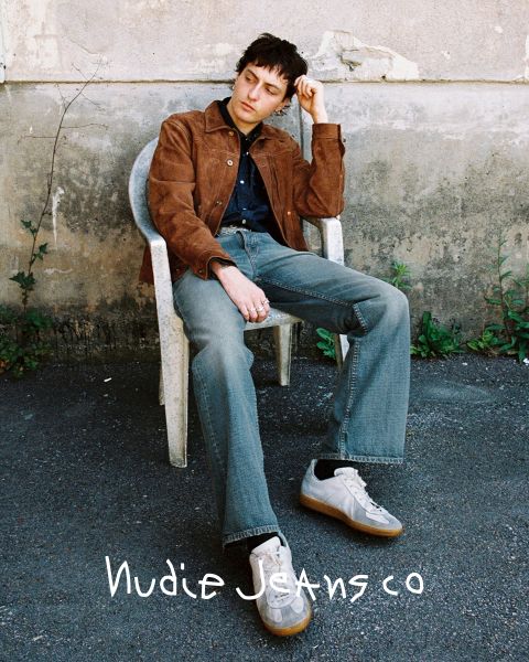 Una persona è seduta all'aperto su una sedia di plastica bianca di fronte a un muro strutturato. Indossa una giacca marrone, jeans e scarpe da ginnastica bianche. In basso si vede il testo "Nudie Jeans Co". Dalla parete crescono erbacce verdi.