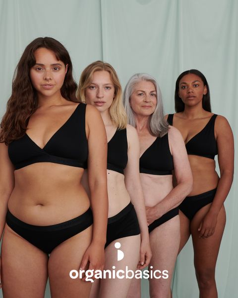 Quattro donne di età e corporatura diverse posano con sicurezza davanti a uno sfondo verde chiaro, indossando biancheria intima nera coordinata. Sul bordo inferiore dell'immagine c'è il testo "organicbasics".