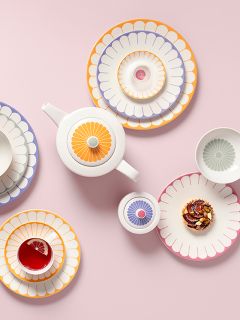 Piatti colorati con motivo floreale, una teiera, una tazza con tè e limone e un pasticcino, disposti su uno sfondo rosa.
