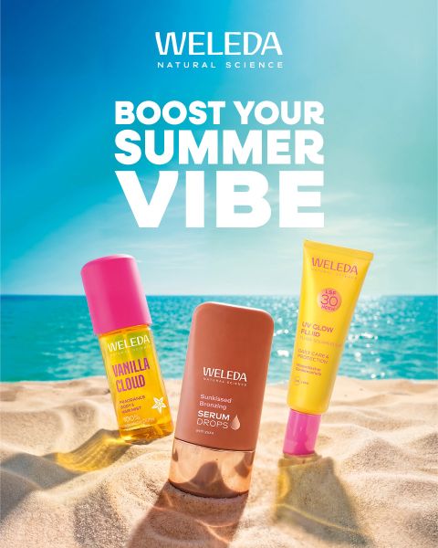 Tre prodotti Weleda per la cura della pelle nella sabbia con lo sfondo del mare e del cielo blu. Testo: "Boost Your Summer Vibe". Scena luminosa e estiva della spiaggia.