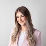 Donna con lunghi capelli castano chiaro che sorride dolcemente, indossa un blazer rosa chiaro sopra un top bianco su uno sfondo bianco semplice.