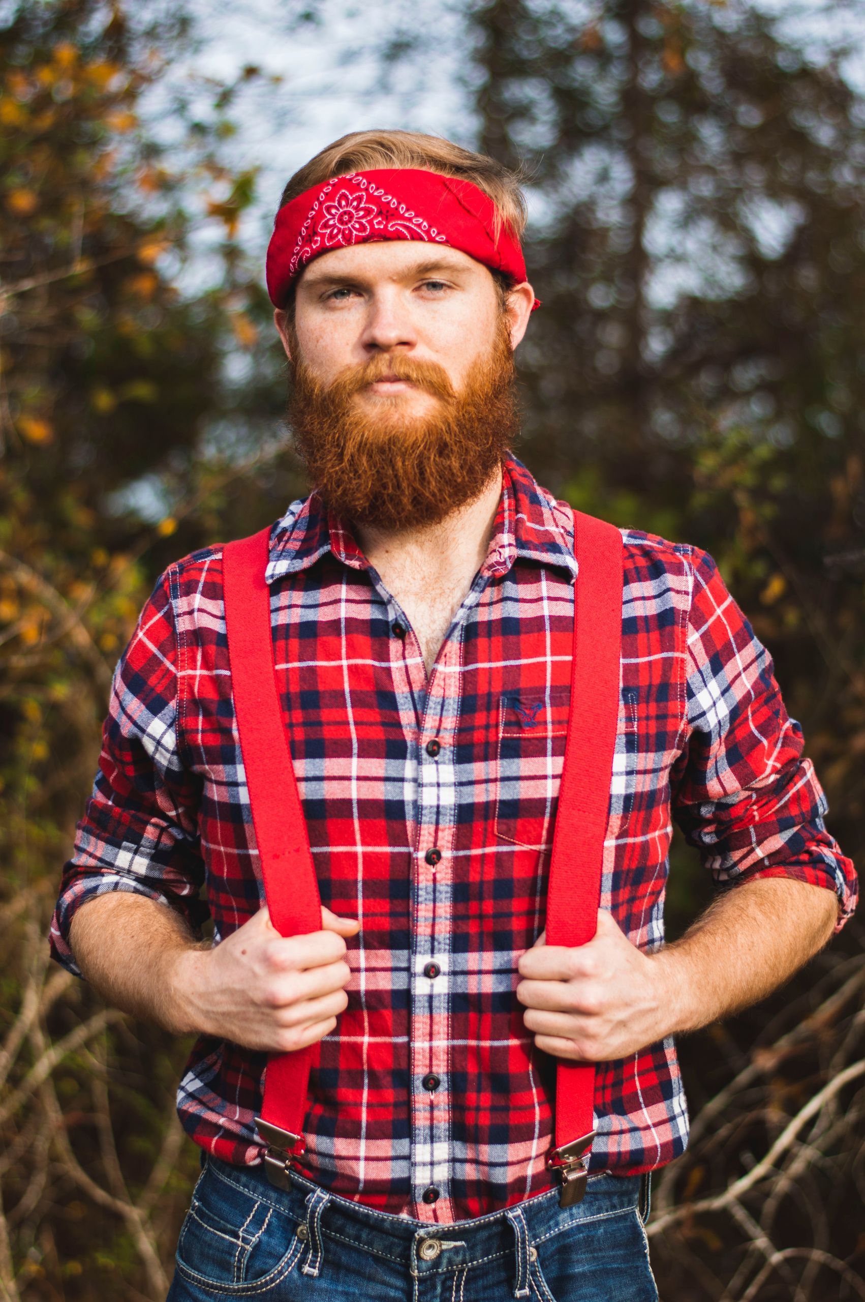 Una persona barbuta con un fazzoletto rosso, una camicia a quadri e bretelle rosse si trova in un ambiente esterno con alberi sullo sfondo.
