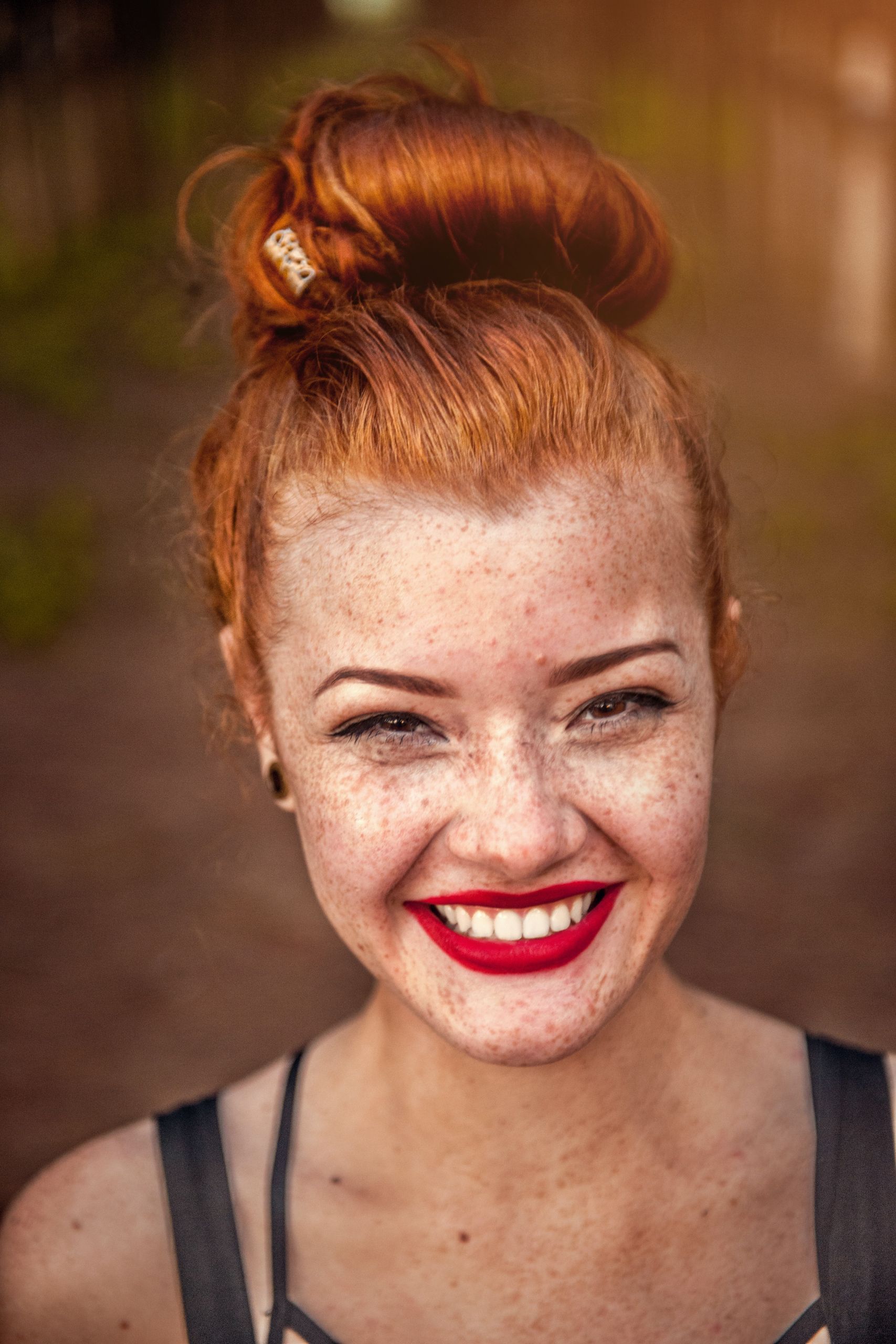 Una donna con i capelli rossi raccolti in uno chignon sorride radiosa, mostrando le sue lentiggini e il suo rossetto rosso. È all'aperto, con uno sfondo sfocato di verde e luce solare.