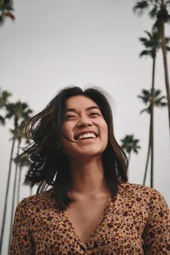 Una persona con i capelli lunghi fino alle spalle è in piedi all'aperto e sorride ampiamente. Sullo sfondo si vedono alte palme davanti a un cielo nuvoloso. La persona indossa un top marrone fantasia.