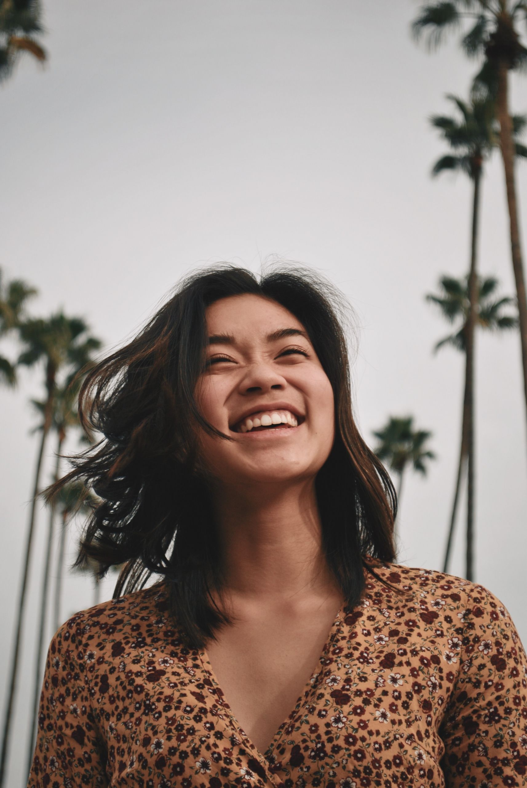 Una persona con i capelli lunghi fino alle spalle è in piedi all'aperto e sorride ampiamente. Sullo sfondo si vedono alte palme davanti a un cielo nuvoloso. La persona indossa un top marrone fantasia.