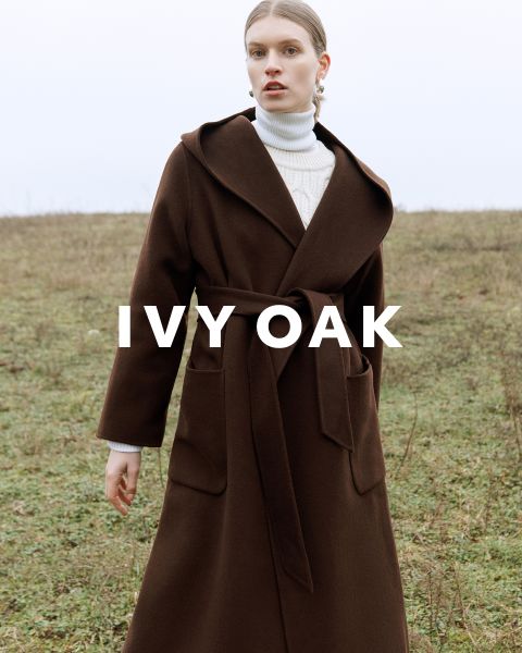 Una persona che indossa un lungo cappotto marrone con cintura è in piedi in un campo aperto. Indossa un dolcevita bianco sotto e guarda avanti. Il testo "IVY OAK" è sovrapposto all'immagine. Lo sfondo è un paesaggio erboso e nuvoloso.