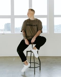 Una persona con i capelli corti è seduta su uno sgabello alto davanti a grandi finestre. Indossa una maglietta "Essentials" marrone, pantaloni neri e scarpe da ginnastica bianche. Sul suo braccio destro si vede un tatuaggio. Lo sfondo è luminoso e minimalista.