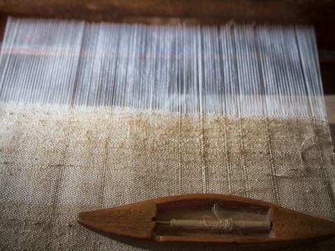 Un telaio fatto a mano con fili parzialmente intrecciati in un tessuto beige. Una navetta di legno si trova sul tessuto, pronta per la tessitura. L'immagine cattura la texture dettagliata dei fili e del tessuto.