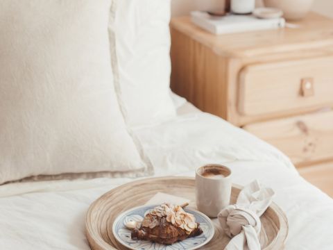 Una scena accogliente in camera da letto con un letto bianco e un comodino in legno. Sul letto, su un vassoio di legno, c'è un piatto con un croissant ricoperto di mandorle, una tazza di caffè e un tovagliolo beige. Sul tavolo ci sono una pila di libri e una candela.