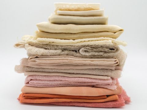 Una pila ordinata di vari tessuti in diverse tonalità di crema, beige, pesca e arancione su uno sfondo bianco semplice. Le texture variano da lisce a leggermente stropicciate.
