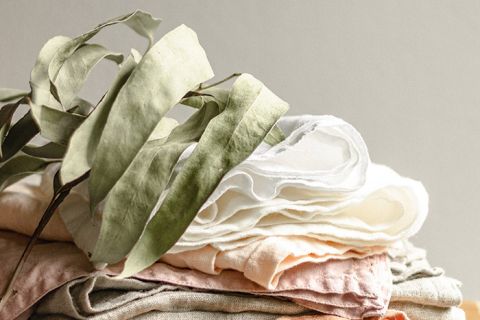 Una pila di tessuti piegati, morbidi e color pastello giace accanto a diverse foglie di eucalipto essiccate, di colore verde chiaro. I tessuti sono nelle tonalità del bianco, crema e rosa chiaro, creando un'estetica tranquilla e naturale.