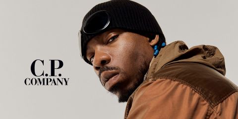Uomo con giacca marrone, berretto nero con occhiali protettivi e perline tra i capelli. Guarda la fotocamera con espressione seria. Sul lato sinistro, su uno sfondo semplice, si trova il testo "C.P. Company".