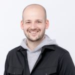 Su uno sfondo bianco semplice, c'è un uomo sorridente con la testa rasata che indossa una giacca nera sopra una felpa con cappuccio grigia.