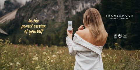 Una donna con lunghi capelli biondi è in piedi in un campo, di spalle, e tiene in mano un prodotto per la cura della pelle. Indossa un top che lascia le spalle scoperte. Il testo recita: "Sii la versione più pura di te stesso". Sullo sfondo si vedono montagne ed erba. Sul display si legge "TRAWENMOOR ORGANIC SKINCARE".