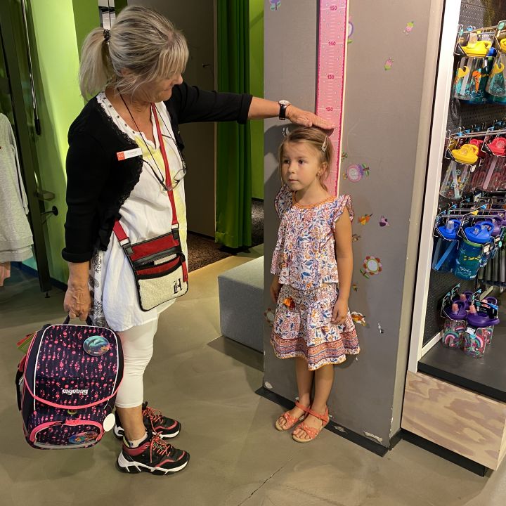 Una donna misura l'altezza di una giovane ragazza usando una bilancia a muro in un negozio. La donna tiene uno zaino colorato e la ragazza è in piedi, indossa un vestito a fiori. Nelle vicinanze, sugli scaffali sono esposte bottiglie d'acqua e vestiti.