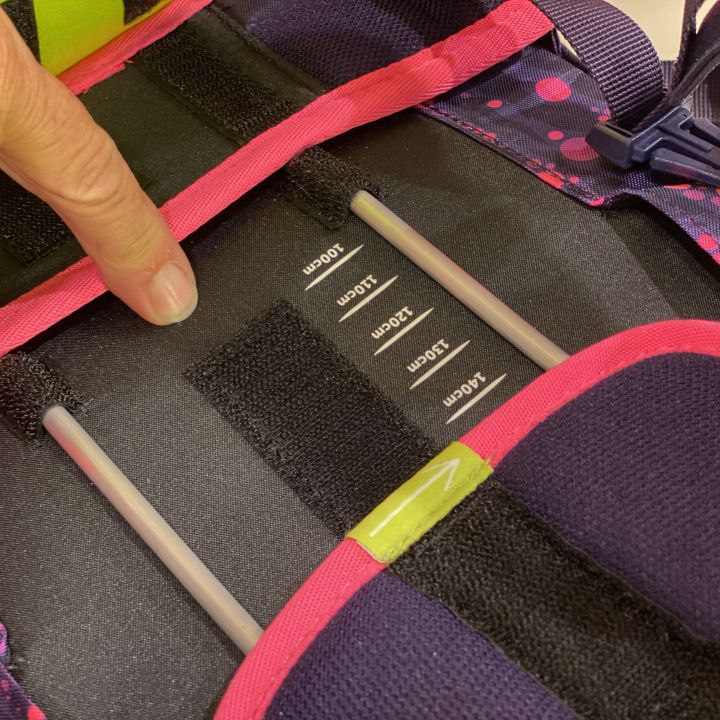 Primo piano della mano di una persona che regola il sistema di cinghie di uno zaino. All'interno ci sono scomparti in velcro con diverse indicazioni di taglia. Lo zaino è progettato nei colori rosa e viola con un motivo a pois.